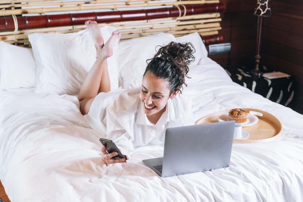 Pretty woman with phone and laptop in bed Huésped disfrutando de conexión WiFi rápida y segura en su habitación de hotel