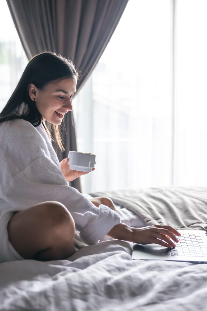 A young woman in a bathrobe works at a laptop in bed.